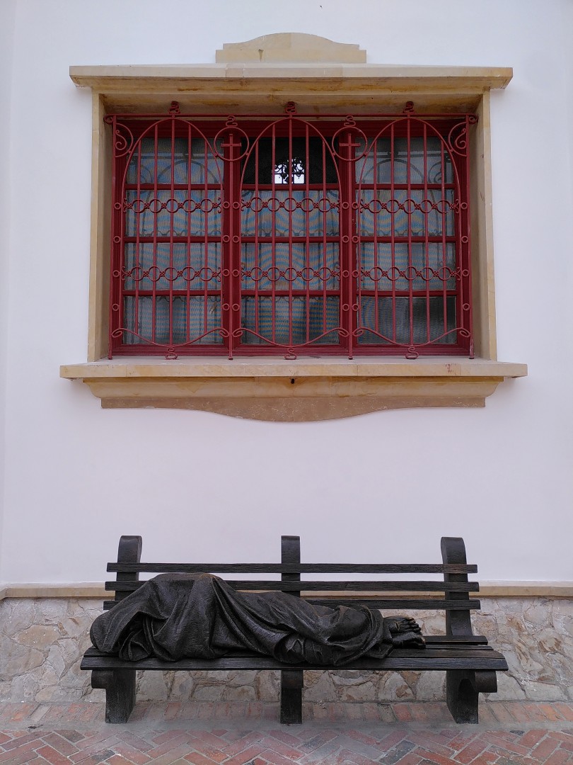 Metal statue of a person with wounded feet, covered by a blanket on a bench in front of a white building