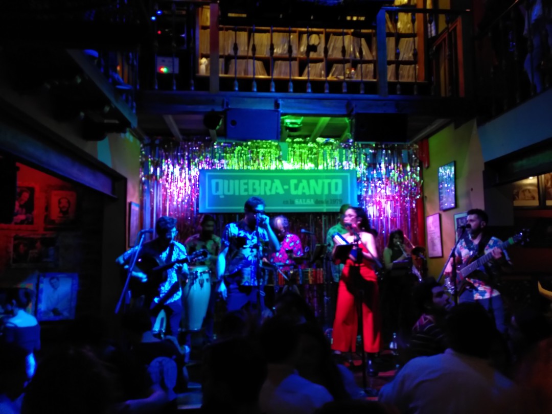 A salsa band of 7 people on stage in a dim room with schemes of a crown in front of it