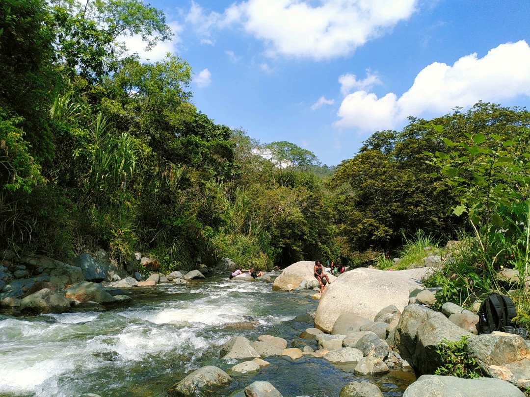 Photo of a river between big rocks and dense green vegetation with people bathing in both sun and water