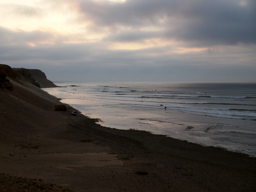 Rocky dunes descending on a flooded sandy beach with waves breaking and the cloudy sky mirroring in the water