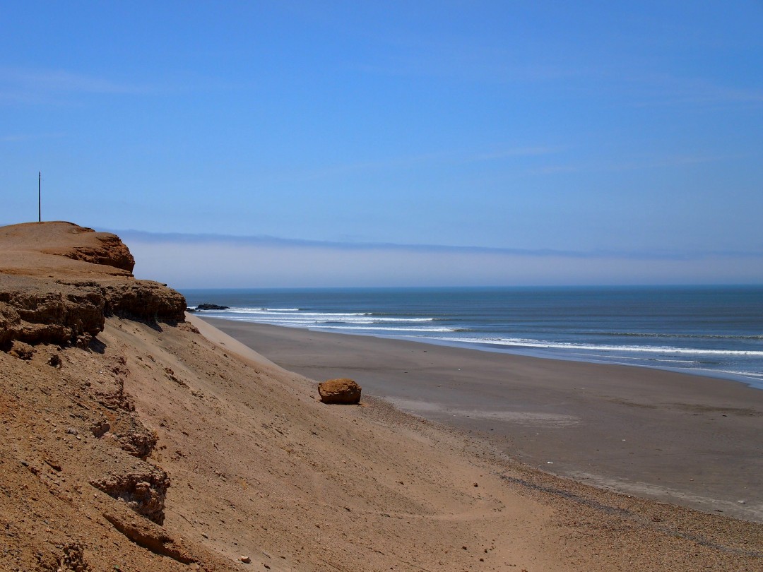 A rocky sand dune descending onto a beach with waves breaking in the distance under a blue sky