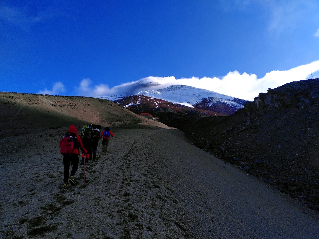 A group of people in colourful, warm outdoor clothing hiking up a sandy trail towards a building at the foot of a massive, snow-capped mountain with its peak in clouds illuminated by the evening sun