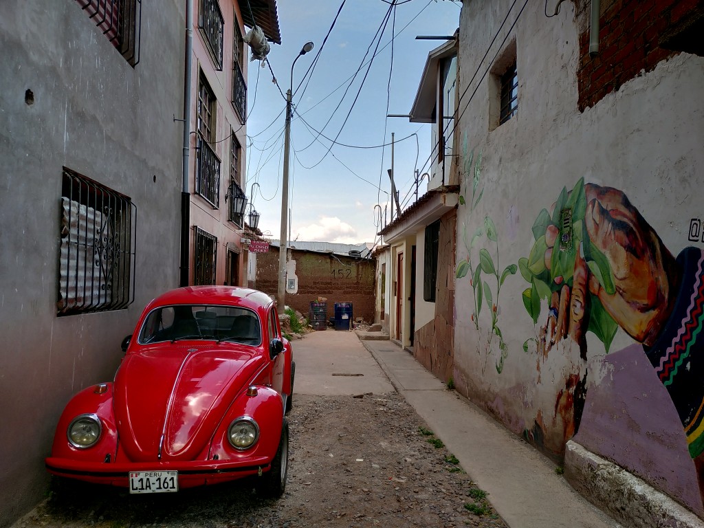 A red VW Beetle parked in a dead end between houses with a graffiti and wires traversing up in the air