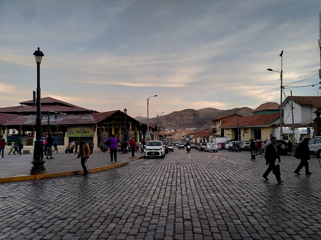 The rusty roof of Cusco's Mercado San Pedro next to a busy cobble-stone street with people and cars