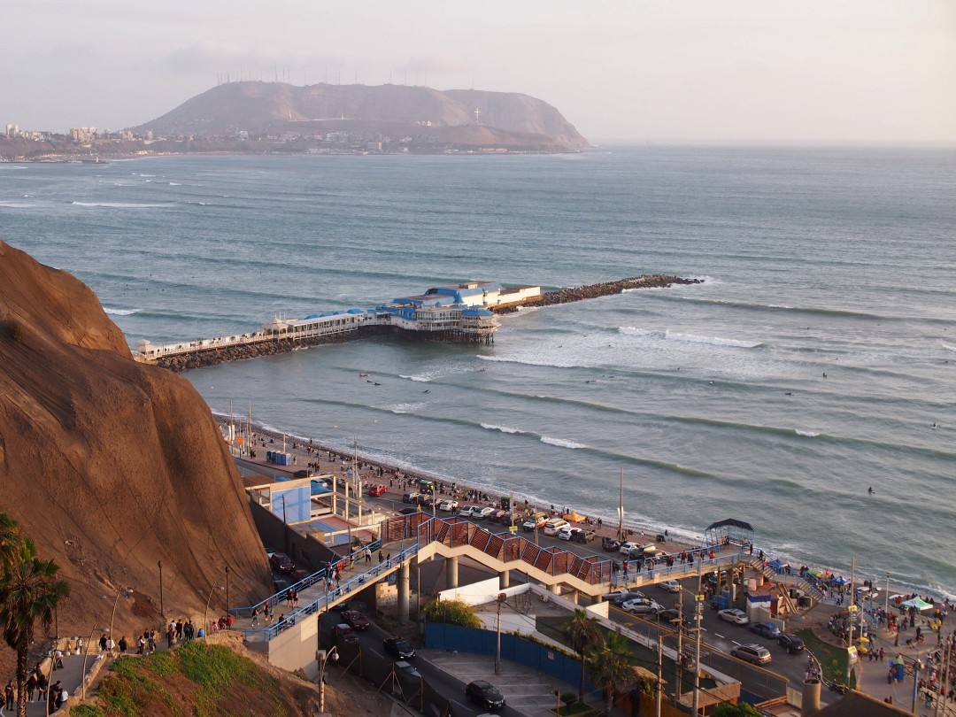 Bird's eye view of Lima's Makaha beach with its stairwell off the cliffs, a restaurant on the nearby jetty and some surfers in the waves