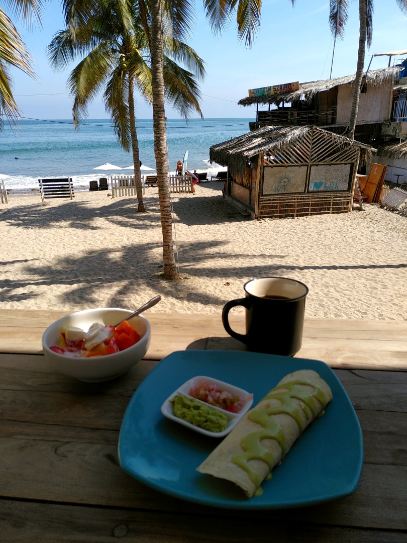 A cup of coffee, some kind of burrito and a bowl of fruit salad on a wooden table with view onto a tropical beach with palm trees and bamboo sheds on it