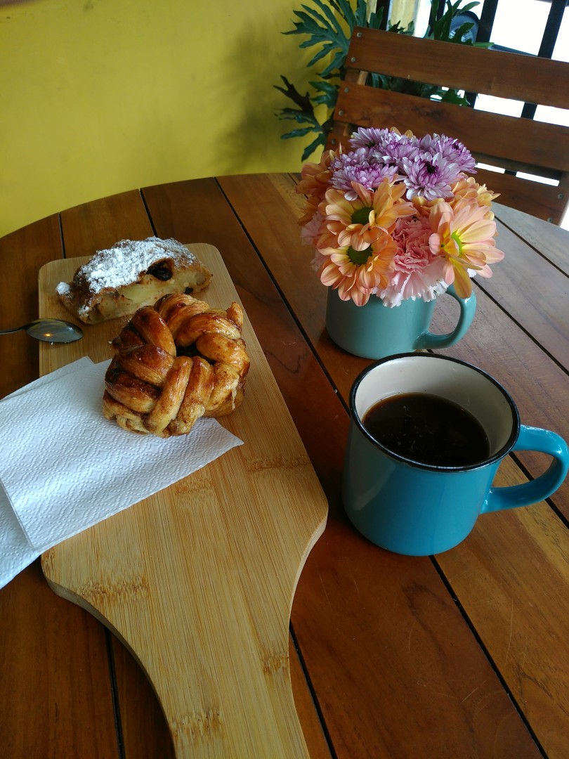 A wooden table with some colourful flowers, a cup of coffee and an equally wooden board with delicious looking pastries on top of it