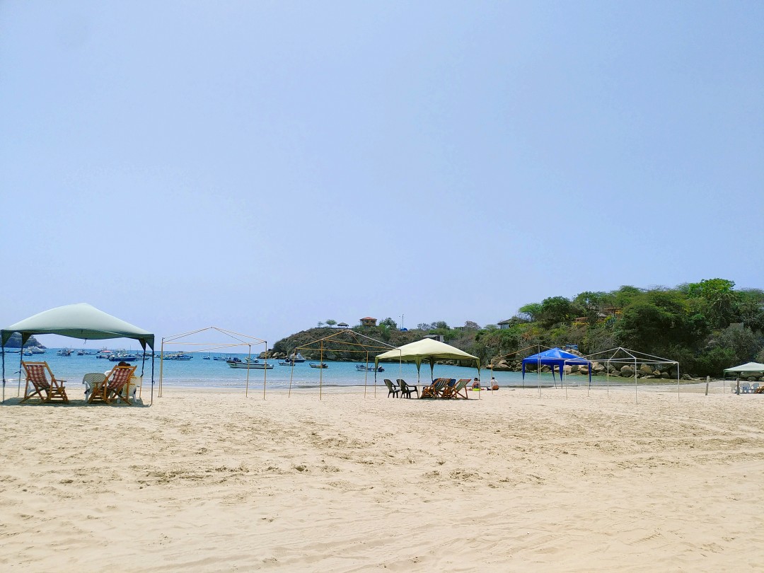 A tropical white beach with some chairs and a bay of light blue water with boats as well as some green trees and bushes at the shore