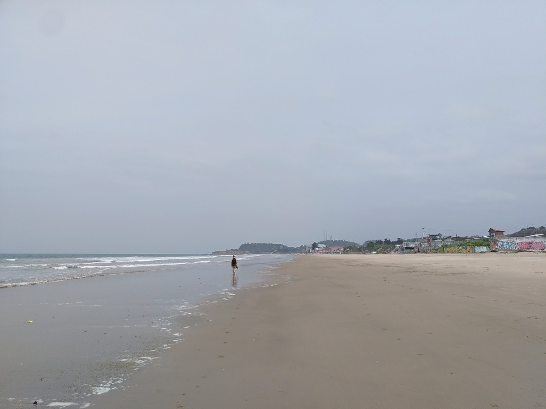 A wide, sandy beach with the ocean to the left, a person walking in the distance and buildings even further in the background