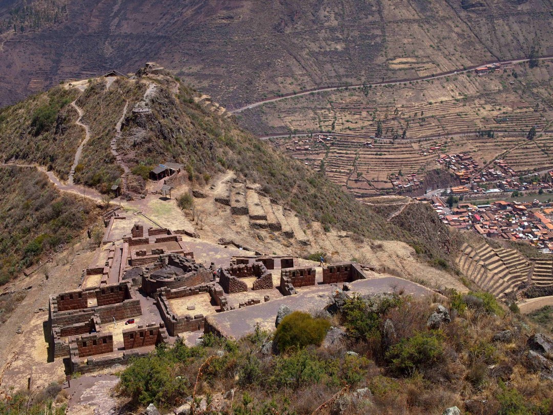 Remains of an Inka temple on a mountain from above with a deep valley and a small contemporary town in the background