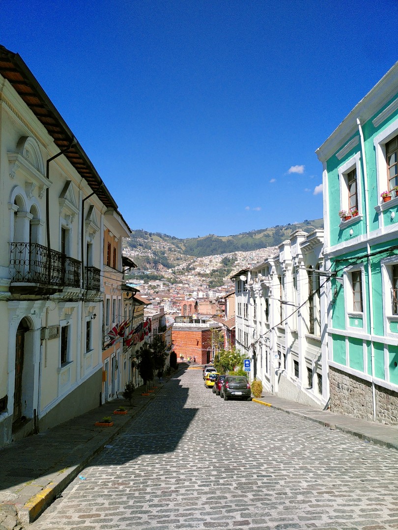 A descending cobblestone street lined with colonial style buildings and a view of the city in the valley and on the opposite mountains