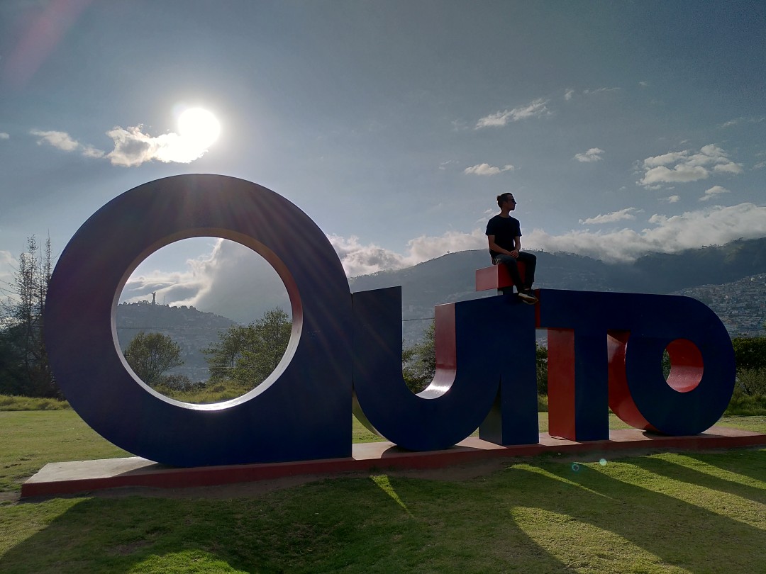 A person sitting on the "i" of big steel letters spelling "Quito", with mountains and the afternoon sun in the background