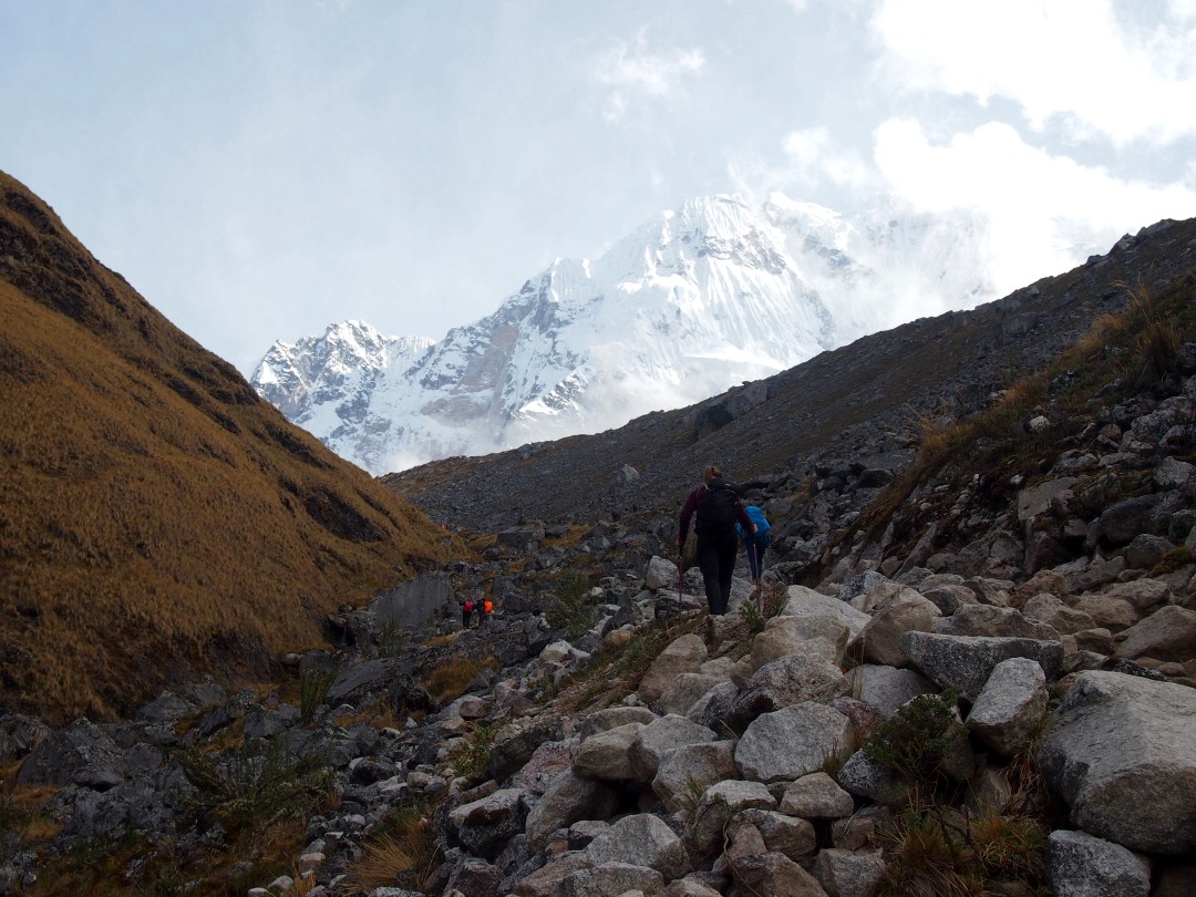 Several people hiking towards snow-covered mountain peeks over rocky terrain