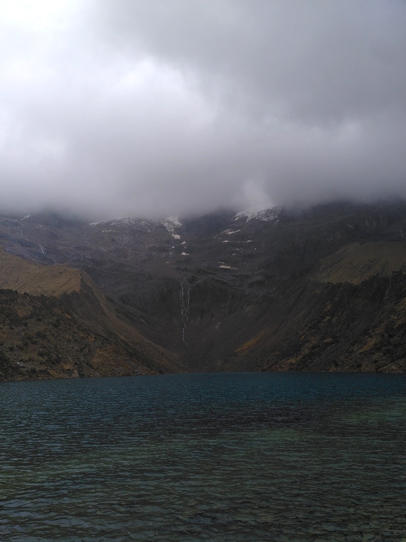 A teal colored lagoon in front of grey mountains that reach into low hanging clouds