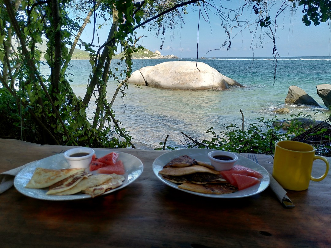 Two plates with pancakes and watermelon as well as a yellow cup of coffee on a wooden table with view of some vegetation and the blue sea behind