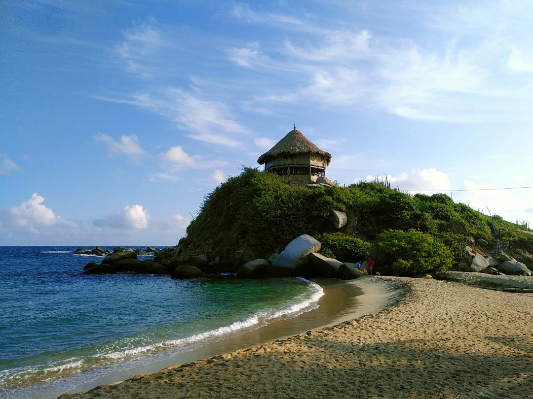 A beach in the morning sun, with blue water, light sand and a wooden house on an overgrown rock