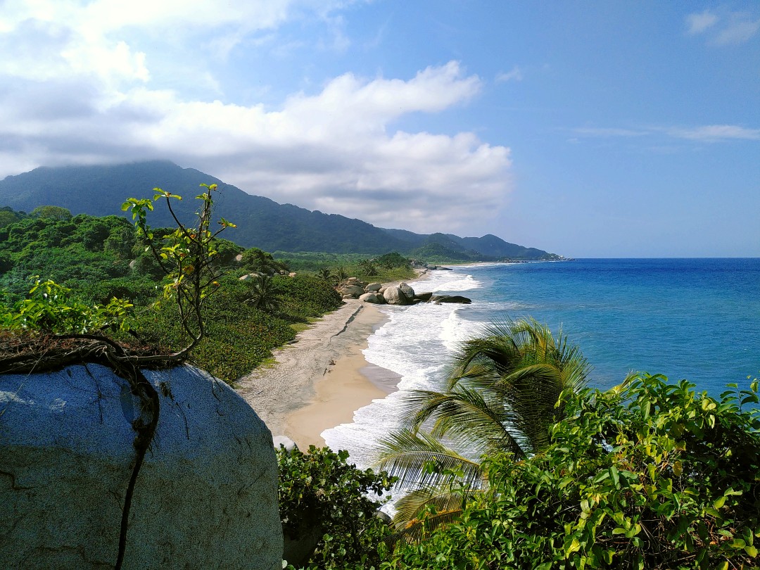 Panorama of a Caribbean coastline with green jungle to the left and blue sea to the right, with some light clouds above